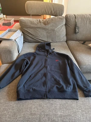 Mörkblå hoodie från Fred Perry - Snygg mörkblå hoodie från Fred Perry med dragkedja framtill och klassisk huva. Tröjan har ribbade muddar vid ärmslut och nederkant samt Fred Perry-loggan broderad i rött på bröstet. Tillverkad i mjuk bomullsblandning, perfekt för en avslappnad stil.
