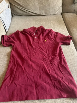 Vinröd pikétröja från Ralph Lauren - Klassisk vinröd pikétröja från Ralph Lauren med korta ärmar och liten gul broderad logga på bröstet. Tröjan har krage och knäppning med två knappar framtill. Perfekt för en stilren och avslappnad look.