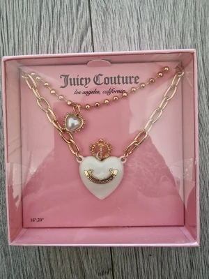 Juicy Couture halsband med hjärthänge - Snyggt halsband från Juicy Couture i guldton med chunky kedja och pärldetaljer. Hängsmycket är ett vitt hjärta med guldfärgad krona och logga, samt en liten pärlprydd hjärtberlock. Perfekt accessoar för att lyfta din outfit.