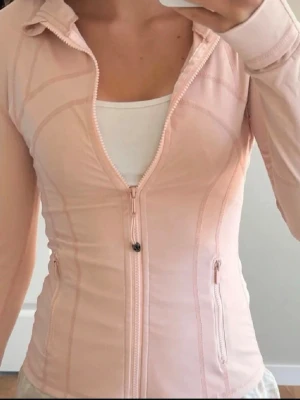 Ljusrosa zip hoodie från Lululemon - Snygg ljusrosa zip hoodie från Lululemon med figurnära passform. Tröjan har långa ärmar, dragkedja framtill och två fickor med dragkedja. Tillverkad i ett stretchigt och mjukt material som passar perfekt till träning eller chill. Strl 4