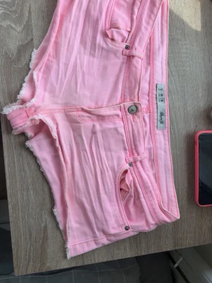 Rosa shorts från Demin Co. - Säljer ett par ljusrosa shorts från Demin Co. med råa kanter och klassisk femficksdesign. De har knapp- och dragkedjestängning samt bälteshällor. Perfekta för varma dagar och ger en fräsch look.
