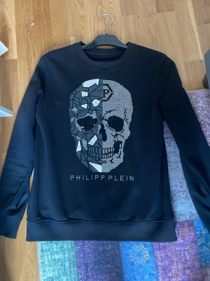 Philipp Plein Sweatshirt - Säljer en svart sweatshirt från Philipp Plein med en stor dödskalle framtill i mosaik och glittrande detaljer. Tröjan har rund halsringning och ribbade muddar vid ärmslut och nederkant. Perfekt för dig som gillar edgy och unik design.