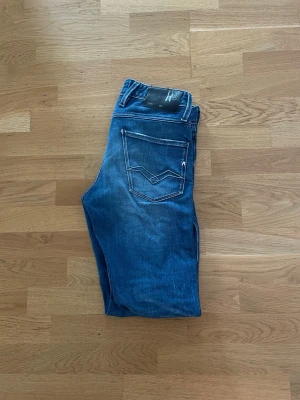 Blå slim fit jeans från Replay - Säljer ett par blå slim fit jeans från Replay med snygga slitningar och klassiska fem fickor. Jeansen har vita kontrastsömmar och en unik bakficka med Replay-logga. Perfekta för dig som gillar en modern och avslappnad stil. Priset kan diskuteras! 