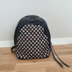 Svart ryggsäck med nitar - Furla - Snygg svart ryggsäck i äkta skinn/ läder & mocka med coola nitar på framsidan från Furla. Väskan har justerbara axelremmar och en dragkedja för säker förvaring. Perfekt för att ge din stil en edgy touch. 34×26cm, botten 25×11cm