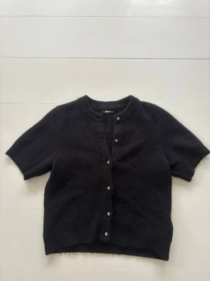 Svart kortärmad kofta Gina Tricot - Kortärmad svart kofta från Gina Tricot med rund halsringning och silvriga knappar framtill. Koftan är stickad i ett mjukt material och har ribbade kanter vid hals och ärmslut. Perfekt att styla över en topp eller klänning.