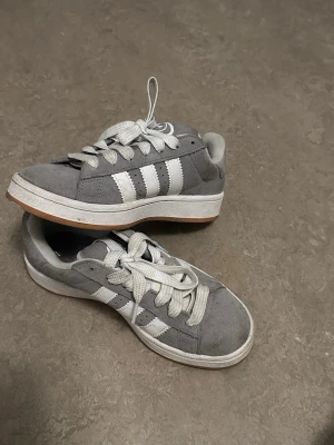Grå Adidas sneakers med vita ränder - Snygga grå sneakers från Adidas med klassiska vita ränder på sidorna och vit sula med brun undersida. Skorna har rund tå, platta snören och är tillverkade i mocka och textil. Perfekt för dig som gillar en clean och sportig look.