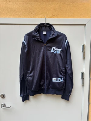 Nike vintage 90s träningsjacka, storlek XL/L - Jackan har vita och ljusblåa ränder på axlarna och ärmarna. På bröstet finns en stor ”C72”-logotyp i ljusblått och vitt. En mindre etikett med text och grafik finns också på vänster sida av jackans nederkant. Baksidan av kragen pryds av texten ”CORTEZ” i vitt. Tröjan är i fint skick. Det som finns att anmärka är löparen på kedjan som syns i sista bilden   Mått  Längd: 68cm  Armhåla till armhåla: 59cm   Har du frågor skicka ett meddelande!   Sökord: vintage , retro , y2k , 00s , fotboll , street
