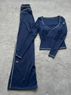 Mörkblått set från Alo med vita detaljer - Snyggt mörkblått set från Alo med långärmad topp och utsvängda byxor. Setet har vita kontrastsömmar längs kanterna och logga på både topp och byxa. Materialet är stretchigt och följsamt, perfekt för en trendig och sportig look.