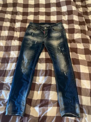 Mörkblå distressed jeans Dsquared2 - Säljer ett par mörkblå jeans från Dsquared2 med slitningar och blekta detaljer. Klassisk femficksmodell med raka ben och låg midja. Jeansen har coola slitningar framtill och en patch med logga bak i midjan. Perfekta för en trendig streetwear-look.