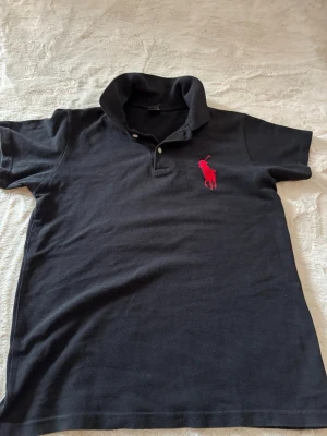 Svart pikétröja från Ralph Lauren - Klassisk svart pikétröja från Ralph Lauren med röd broderad logga på bröstet. Tröjan har krage, korta ärmar och knappar vid halsen. Perfekt till jeans eller shorts för en clean och tidlös look.