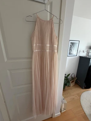 Lång ljusrosa klänning med tyll - Elegant långklänning i ljusrosa med tunna axelband och vackert fallande tylltyg. Klänningen har markerad midja med diskreta banddetaljer och en luftig, svepande kjol. Perfekt för speciella tillfällen och ger en romantisk känsla.