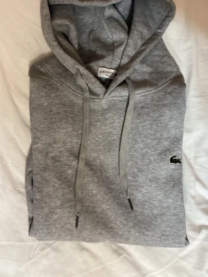 Grå hoodie från Lacoste - Snygg grå hoodie från Lacoste med klassisk krokodillogga på bröstet. Hoodien har dragsko i huvan och är gjord i mjukt material som känns skönt mot huden. Perfekt för en chill och stilren look.