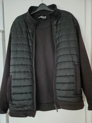 Cardiganjacka - Stilren svart cardiganjacka med quiltad framsida och mjuka ärmar. Jackan har hög krage, dragkedja framtill och två fickor. Perfekt för dig som gillar en enkel och modern look.