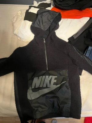 Svart Nike hoodie med stor logga - Säljer en svart hoodie från Nike med halv dragkedja och stor ficka framtill med tydlig Nike-logga i grått. Huvan är i ett glansigt material medan resten av hoodien är i mjuk teddyfleece. Perfekt för en sportig och avslappnad stil.