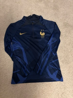 Blå Nike Frankrike träningströja - Mörkblå långärmad träningströja från Nike med Frankrikes landslagslogga och två stjärnor i guld på bröstet. Tröjan har halv dragkedja framtill och ett grafiskt mönster i blått och guld. Perfekt för fotbollsträning eller sportiga aktiviteter.
