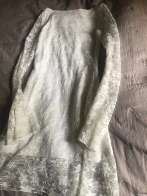 Vit spetsklänning med lång ärm - Säljer en vit klänning i spets med långärmad design och blommigt mönster. Klänningen har en rak passform och är kort i modellen. Perfekt för dig som gillar romantisk och feminin stil.