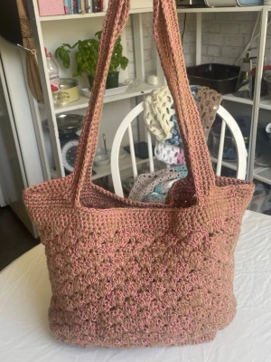Handvirkad shoppingväska i rosa och beige - Unik handvirkad shoppingväska i mjukt garn med mix av rosa och beige toner. Väskan har två rejäla axelband och ett snyggt, strukturerat mönster. Perfekt för dig vill ha något personligt.