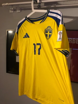 Svensk landslagströja Adidas gul - Gul svensk landslagströja från Adidas med blå detaljer och nummer 17. Tryck med 'GYÖKERES' på ryggen och officiella FIFA-märken på ärmarna. Klassisk rund hals och korta ärmar, perfekt för fotbollsfans.