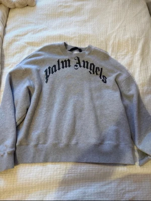 Grå sweatshirt från Palm Angels - ÄKTA - Snygg grå hoodie från Palm Angels med svart logga i gotisk stil över bröstet. Oversized passform och ribbade muddar vid ärmslut och midja. Tillverkad i mjukt bomullsmaterial, perfekt för en avslappnad streetwear-look. , liten defekt vid A som man ser på bilden. NYPRIS - 2800 kr  