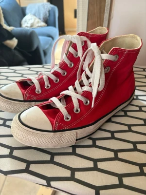 Röda Converse Chuck Taylor High - Klassiska röda Converse Chuck Taylor All Star high tops med vit tå och sula. Skorna har vita snören, metallöljetter och den ikoniska loggan på sidan. Tillverkade i slitstark canvas med gummisula. Perfekta för en cool och avslappnad stil.