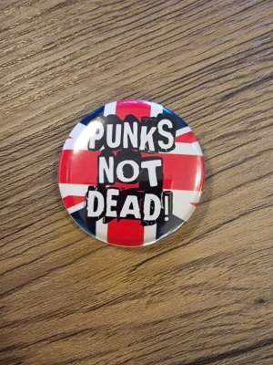 Punks Not Dead pinsknapp - Cool knapp med texten 'Punks Not Dead' i svart graffiti-stil. 4,4 cm. Perfekt accessoar för dig som gillar punk och vill sticka ut. Rund form och klassisk punkkänsla.