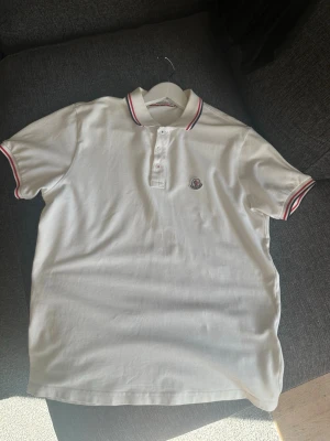 Moncler - Vit pikétröja från med klassisk krage och knappar framtill. Tröjan har röda och blå ränder på krage och ärmslut samt logga på bröstet. Tillverkad i bomull och har en normal passform med korta ärmar.