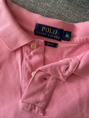 Rosa pikétröja från Polo Ralph Lauren - Säljer en klassisk rosa pikétröja från Polo Ralph Lauren i storlek XL. Tröjan har krage, knappar framtill och den ikoniska blå broderade loggan på bröstet. Tillverkad i bomull med en skön och luftig känsla. Perfekt för en avslappnad men snygg stil.