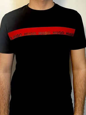 Svart HUGO t-shirt med röd logga - Snygg svart t-shirt från HUGO med en bred röd rand över bröstet där HUGO-loggan upprepas i svart text. Klassisk passform och rund halsringning. Perfekt för dig som gillar stilrena plagg med en tydlig logga. ÄKTA VARA! 