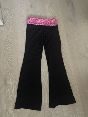 Svarta yogapants med rosa leopardmönster - Säljer ett par svarta yogapants med utsvängda ben och en bred midjeresår i rosa med leopardmönster. Materialet känns stretchigt och bekvämt, perfekt för en avslappnad och trendig look. 