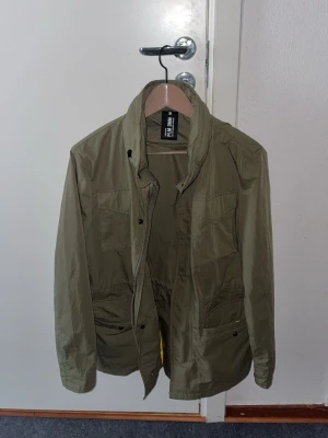 Olivgrön vindjacka från Pull & Bear - Snygg olivgrön vindjacka från Pull & Bear med hög krage och flera praktiska fickor framtill. Jackan har dragkedja och tryckknappar samt justerbar midja för bättre passform. Perfekt för dig som gillar enkel och stilren design.