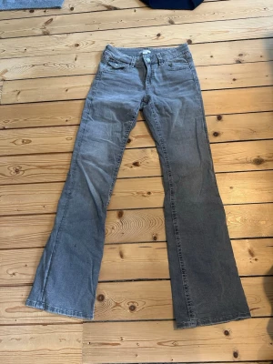 Grå bootcut jeans med fickdetaljer - Snygga grå bootcut jeans med klassiska fem fickor och dekorativa ficklock bak. Jeansen har låg midja och vida ben, perfekta för en avslappnad och trendig look. Tillverkade i mjukt denimtyg som ger en skön passform.