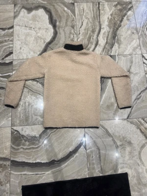 Beige teddytröja från Snoot - Mysig beige teddytröja från Snoot med svarta detaljer vid krage, ärmslut och nederkant. Tröjan har en rak passform och är tillverkad i ett fluffigt, fårskinnsliknande material som håller dig varm under kyliga dagar.
