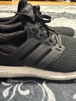 Svarta Adidas Ultraboost sneakers - Säljer ett par svarta Adidas Ultraboost sneakers med stickad ovandel och klassiska tre ränder på sidan. Skorna har vita Boost-sulor för extra komfort och svarta skosnören. Perfekta för dig som gillar stilrena och sportiga sneakers. Ny utan box 