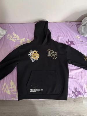 Svart hoodie med tryck från Silly Dino - Svart hoodie från Silly Dino med stor huva och magficka. Framtill finns coola tryck i vitt och gult, bland annat ett S och NY i graffiti-stil. Tryck även på ena ärmen. Tillverkad i mjukt material, perfekt för en streetwear-look.