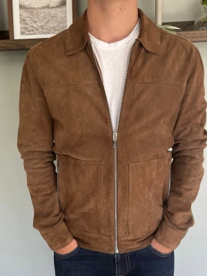 Reiss Mocka Jacka – 100% Getskinn | Cognac/Tan | Strl Xs - REISS MOCKA JACKA  Storlek: XS Passform: Slim/normal Färg: Cognac/tan Material: 100% getskinn (mocka) Foder: 100% polyester Tillverkad i Indien  En riktig investeringsjakka från brittiska Reiss. Heltäckande dragkedja i silver, två zipfickor i fronten och en klassisk skjortkrage. Mjukt getskinn med en naturlig textur som bara blir bättre med åren.  Passar perfekt till jeans, chinos eller mörka byxor — en jacket som lyfter vilket outfit som helst.  Modellen på bilden är 175 cm och väger 65 kg.  
