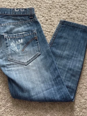Blå slitna jeans dondup - Säljer ett par blå jeans med snyggt sliten look och patchdetalj på bakfickan. Jeansen har klassisk femficksmodell och raka ben. Perfekta för en avslappnad och trendig stil. I storlek 32