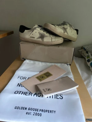 Golden Goose vita sneakers med stjärna - Golden Goose sneakers i vitt läder med grå snörning och ikonisk silverfärgad stjärna på sidan. Skorna har svart detalj vid hälen och klassisk låg siluett. Kommer med originalkartong, dustbag och äkthetskort. Perfekta för en trendig och avslappnad stil.