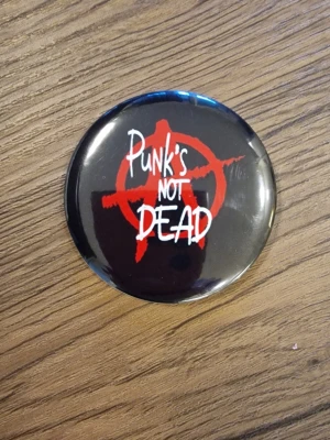 Punks Not Dead pinsknapp - Cool knapp med texten 'Punks Not Dead' i svart graffiti-stil. 5,8 cm. Perfekt accessoar för dig som gillar punk och vill sticka ut. Rund form och klassisk punkkänsla.