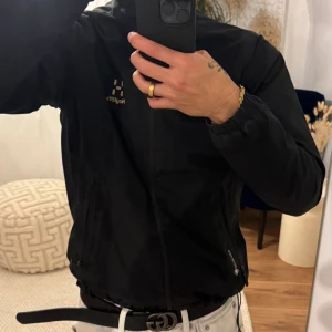 Svart vindjacka från Haglöfs - Svart vindjacka från Haglöfs med huva och dragkedja framtill. Jackan är tillverkad i lätt och slitstark polyester med meshfoder på insidan. Snygg logga på bröstet och Core-Tex-detalj vid fickan. Perfekt för dig som vill ha en clean och sportig look. Jackan har en liten defekt på en av fickorna gällande kedjan. 