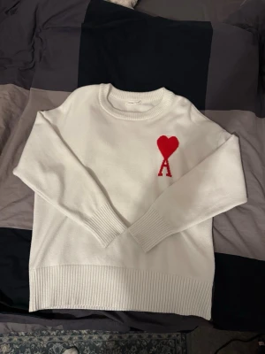 Vit stickad tröja Ami Paris hjärta - Säljer en vit stickad tröja från Ami Paris med röd hjärtlogga och bokstaven A på bröstet. Tröjan har rund halsringning, långa ärmar och ribbade muddar vid ärmslut och nederkant. Perfekt för dig som gillar stilrena plagg med en twist.