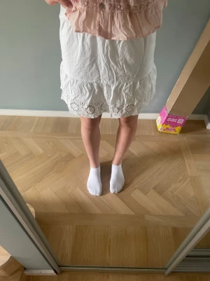 Vit kjol med broderad volangkant - Söt vit kjol med bred volangkant och broderade detaljer längst ner. Kjolen har en avslappnad passform och är perfekt för en somrig look. Tillverkad i lätt bomullstyg som känns luftigt och bekvämt.
