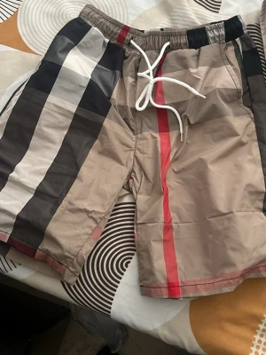 burberry  - Snygga shorts med breda ränder i beige, svart, vitt och rött. De har elastisk midja med vit snörning och en avslappnad passform. Perfekta för sommaren och ger en trendig look med sitt klassiska mönster.