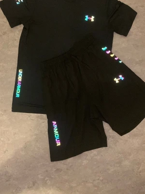 Svart set från Under Armour - Snyggt svart set från Under Armour med t-shirt och shorts. Båda plaggen har regnbågsfärgade loggor och textdetaljer. Materialet är mjukt och stretchigt, perfekt för en sportig look. Passar dig som gillar färgglada detaljer och bekväma kläder.