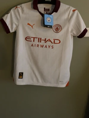 Manchester City bortatröja Foden 47 - Snygg Manchester City bortatröja från Puma med Foden 47-tryck på ryggen. Tröjan är vit med vinröda detaljer på krage och ärmslut, samt orange Puma-logga och Etihad Airways sponsortryck. Tillverkad i lätt och ventilerande material, perfekt för fotbollsträning eller match.