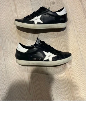 Golden Goose Super-Star sneakers svart/vit - Golden Goose Super-Star sneakers i svart och vitt läder med klassisk låg silhuett. Skorna har svart yttersula med fiskbensmönster och tydlig logga på innersulan. Perfekta för dig som gillar streetstyle och vill ha en unik touch på outfiten. Lite små i storleken. Säljer då de var för små, köpa här på pick.