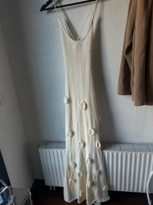 Lång vit stickad klänning med blommor - Lång vit stickad klänning med smala axelband och virkade blommor som dekoration längs kjolen. Klänningen har en figurnära passform och djup v-ringning. Klänningen har öppen rygg som knyts bak själv 💕💕aldrig använd, storlek s