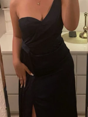 Oneshoulder balklänning - Snygg marinblå långklänning med one shoulder-design och omlott framtill. Klänningen har en markerad midja och en slits som ger en elegant touch. Perfekt för dig som vill sticka ut på bal eller fest. Materialet känns mjukt och följsamt.