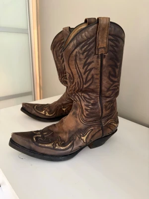 Bruna cowboy boots med brodyr - Säljer ett par klassiska cowboy boots i brunt läder med snygga broderade detaljer i svart och guld. Bootsens spetsiga tå och höga skaft ger en autentisk westernkänsla. Perfekta för dig som vill sticka ut med en cool och tidlös stil.