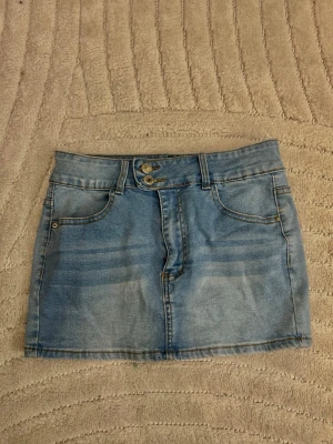 Ljusblå jeanskjol med dubbla knappar - Trendig ljusblå jeanskjol i kort modell med dubbla knappar framtill och klassiska fickor både fram och bak. Kjolen har bälteshällor och en snygg tvättad denimlook som passar perfekt till vardagsoutfits.