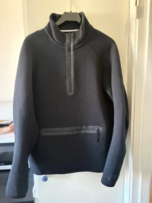 Svart half zip Nike tech hoodie med dragkedja - Snygg svart half zip från Nike med hög krage och halv dragkedja framtill. Praktisk ficka med dragkedja på magen och sportig design. Tillverkad i mjukt och bekvämt material som passar perfekt till streetwear eller chill dagar.